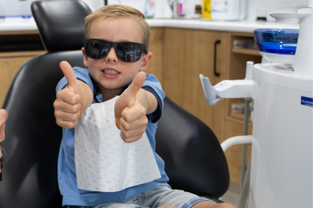 Boy in a dental clinic giving two thumbs up, wearing sunglasses and a patterned bib in a dental chair.