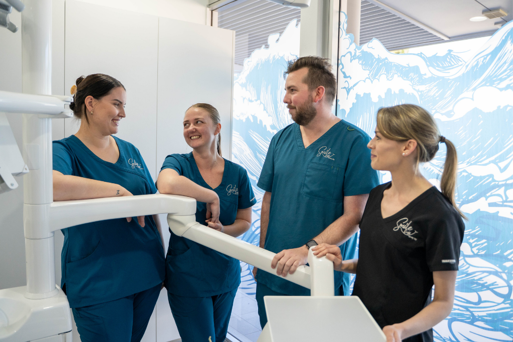 Four healthcare professionals in teal scrubs stand together in a bright dental clinic, smiling near imaging equipment.