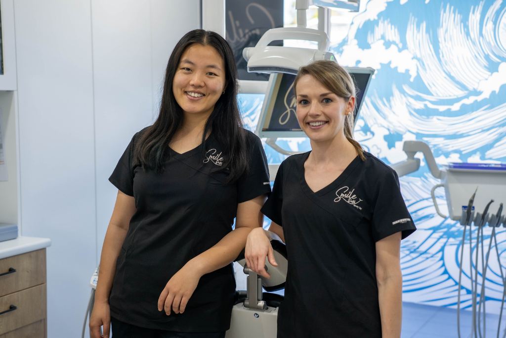 Two dental assistants in black scrubs smile at the camera beside a dental chair in a modern clinic.