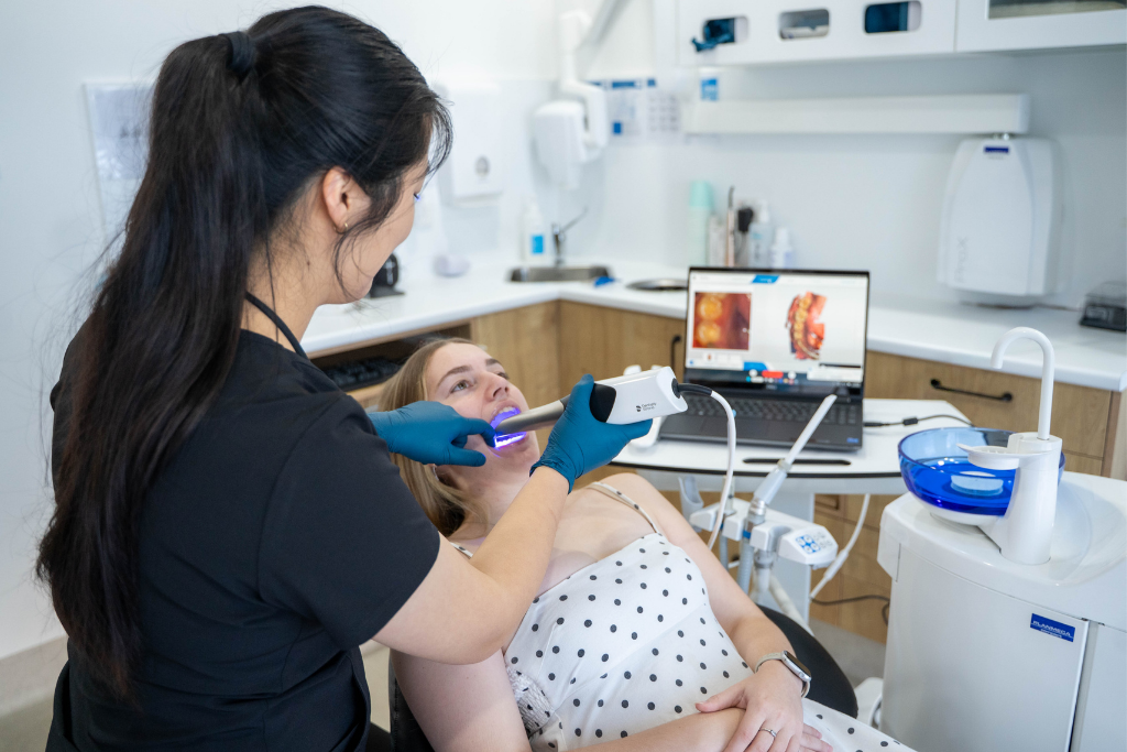 Dental professional in blue gloves using a curing/light device on a patient in a dental chair, laptop showing dental images nearby.